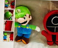 Plush toy resembling a character in green hat and blue overalls next to a red and black plush toy on a textured surface.