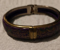 A close-up of a hinged bangle bracelet with a purple and gold patterned inlay and a silver-toned metal clasp, resting on a textured white surface.