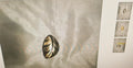 A metallic oval pin with a leaf-like cutout pattern in a gold and silver-tone finish, displayed on a textured light gray fabric background with three smaller thumbnail views on the right.